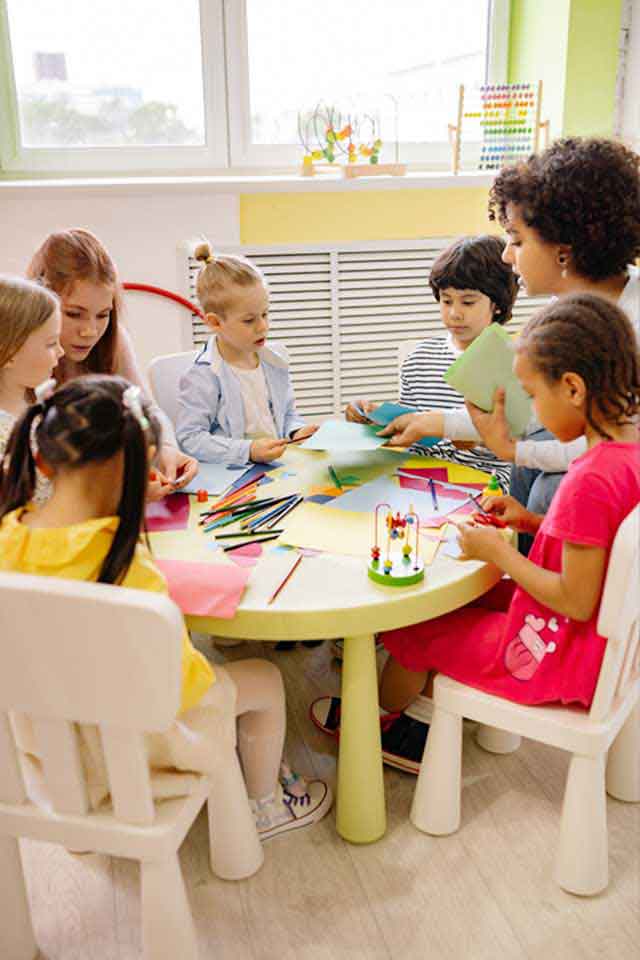 Children sitting on chairs in front of her teacher on roundtable at classroom doing art